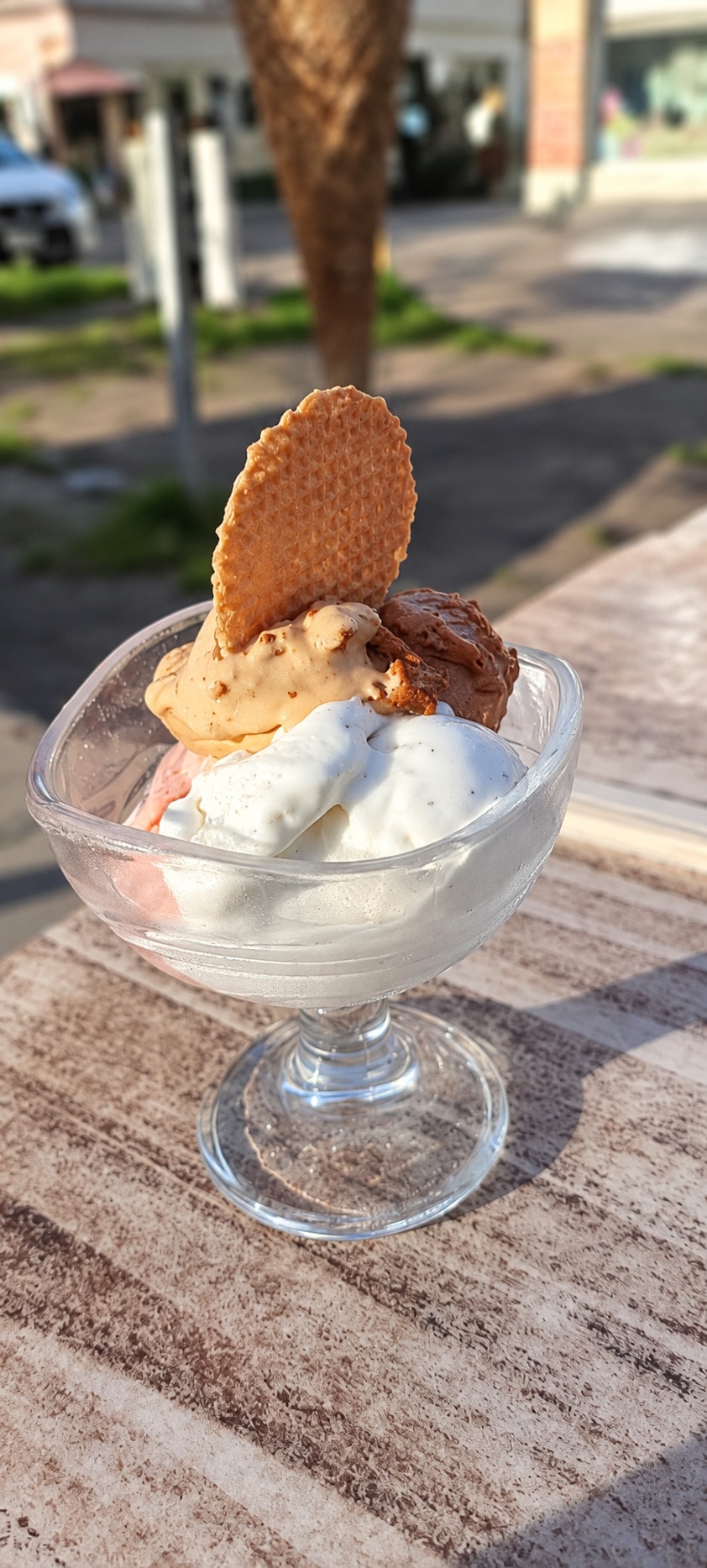 A glass coupe of vanilla and caramel gelato with a wafer on a sunny table outside