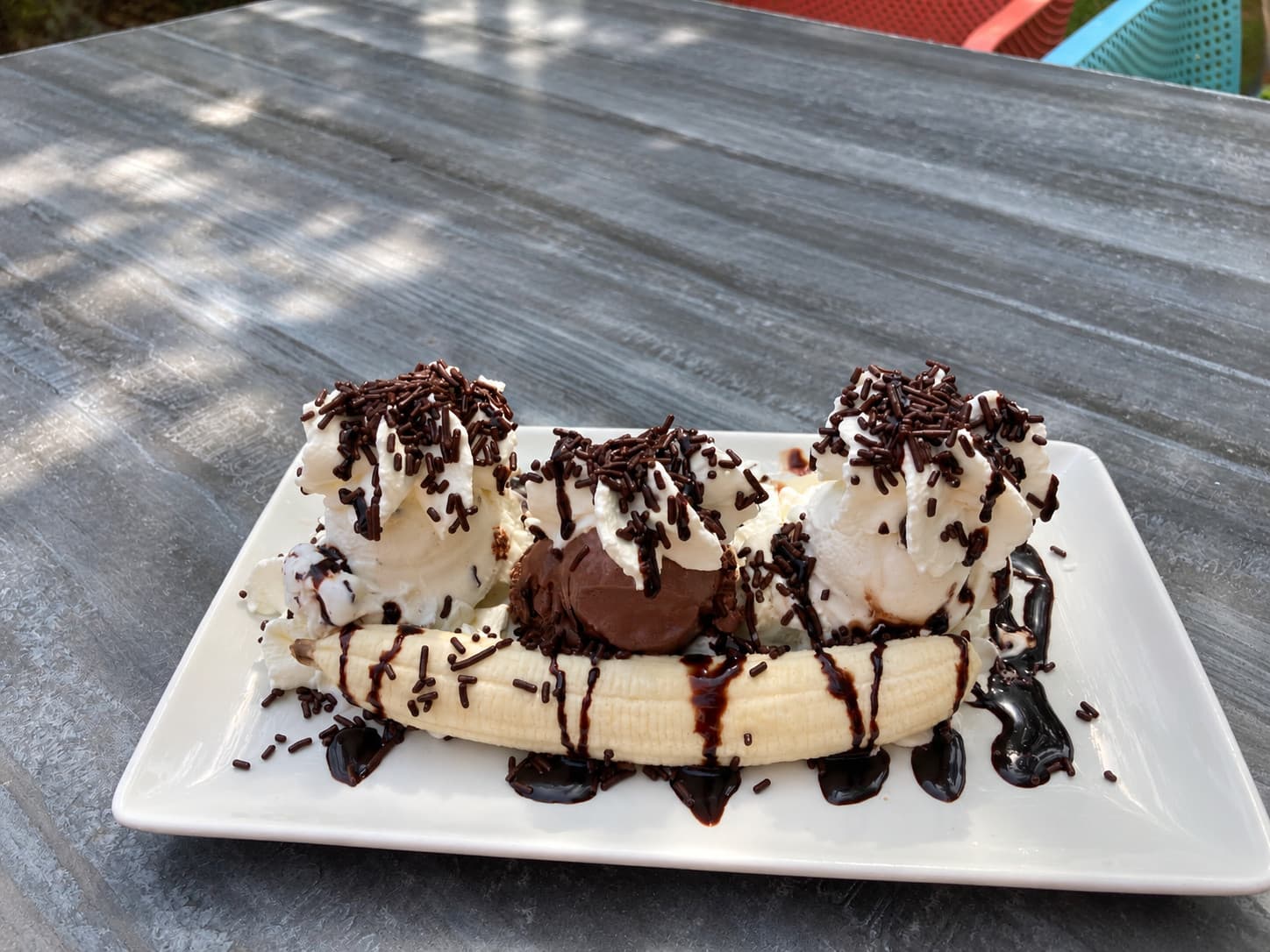 A banana split with three scoops, cream, chocolate sauce and sprinkles on a square plate