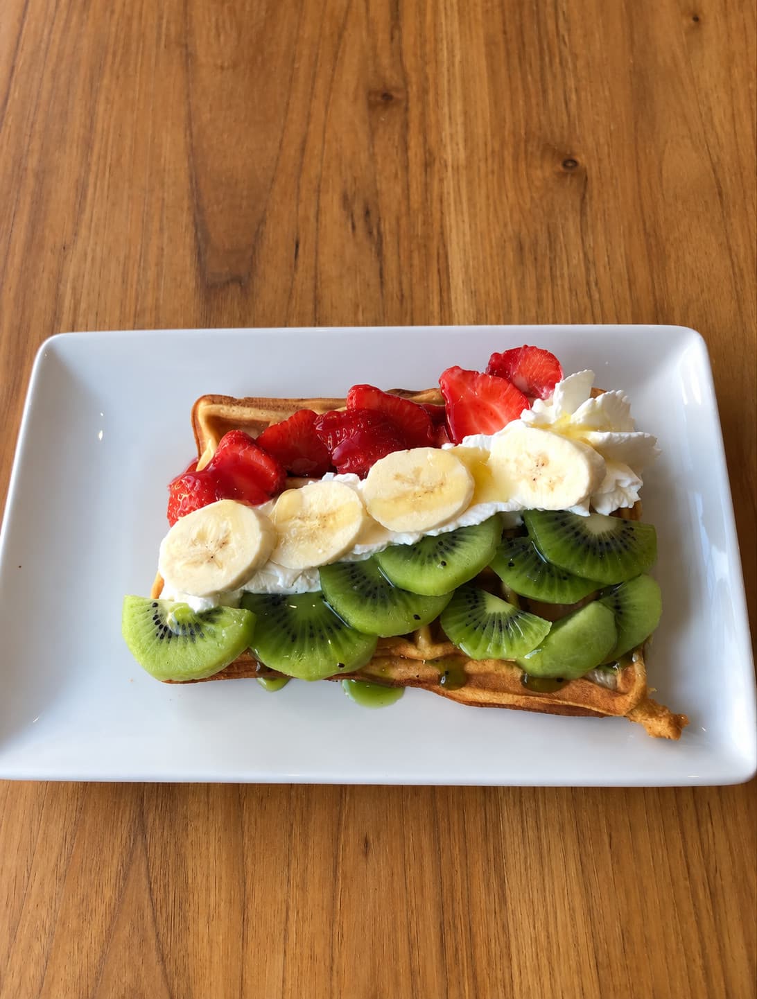 A Belgian waffle topped with strawberries, banana, kiwi and cream on a white plate