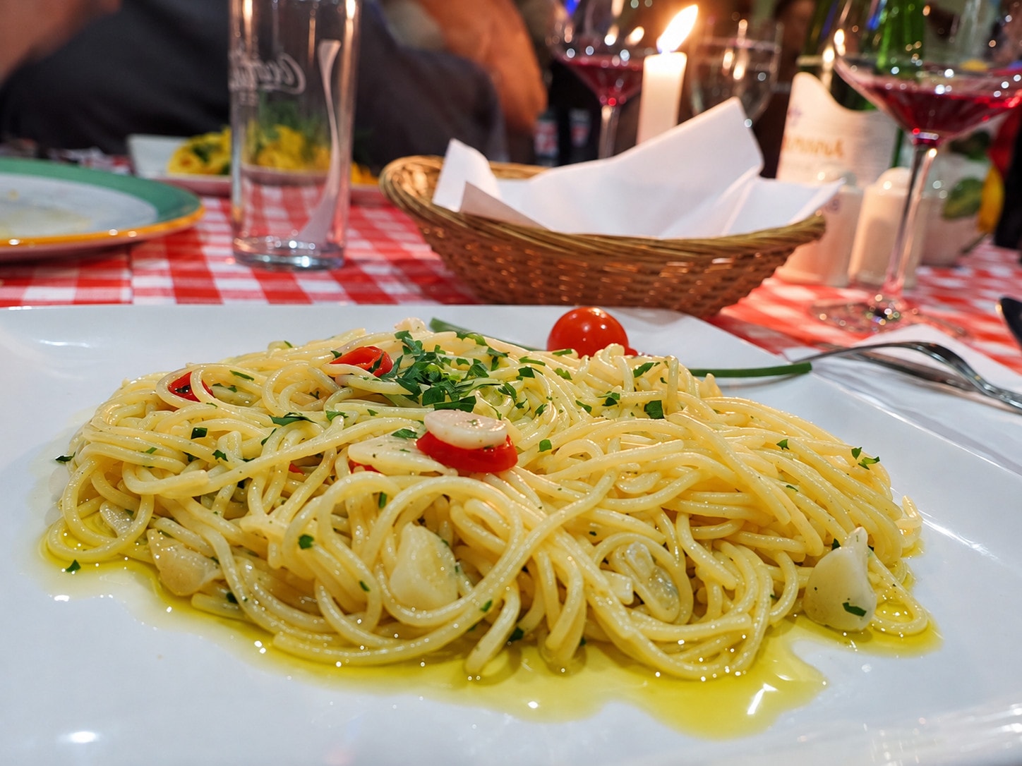 A plate of spaghetti by candlelight with wine glasses and a bread basket behind